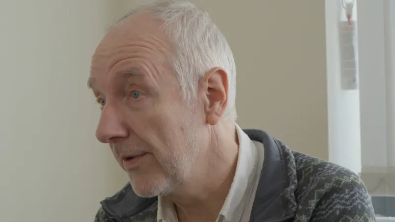 A Still of Dr Phil Davison Side on talking in a interview against a beige wall.